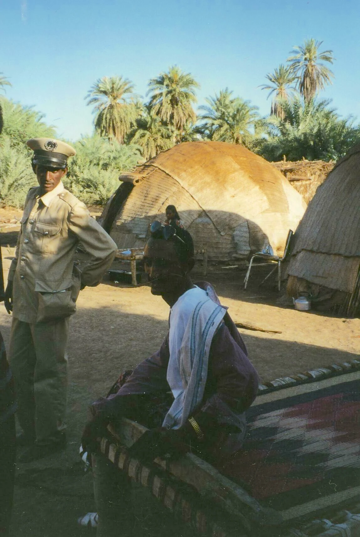 Mohammed sits on a woven bed in an Afar village while a policeman looks towards the camera